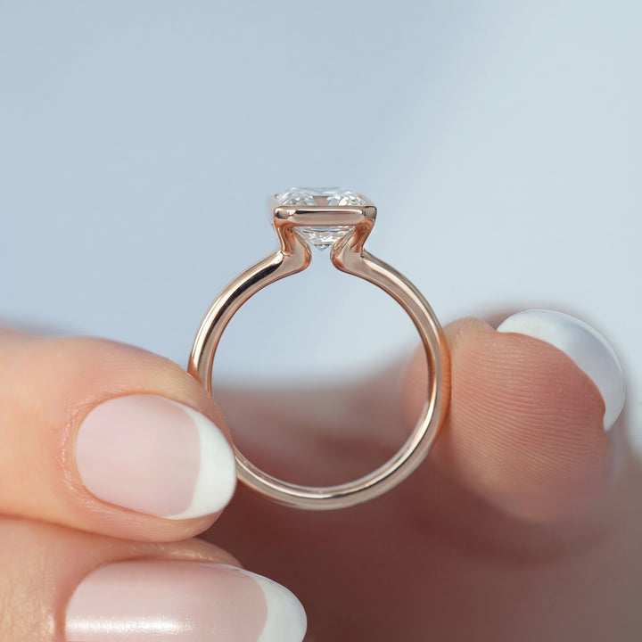 A close-up of a rose gold solitaire ring with a clear rectangular gemstone held between fingers against a soft light blue background.#material_rose