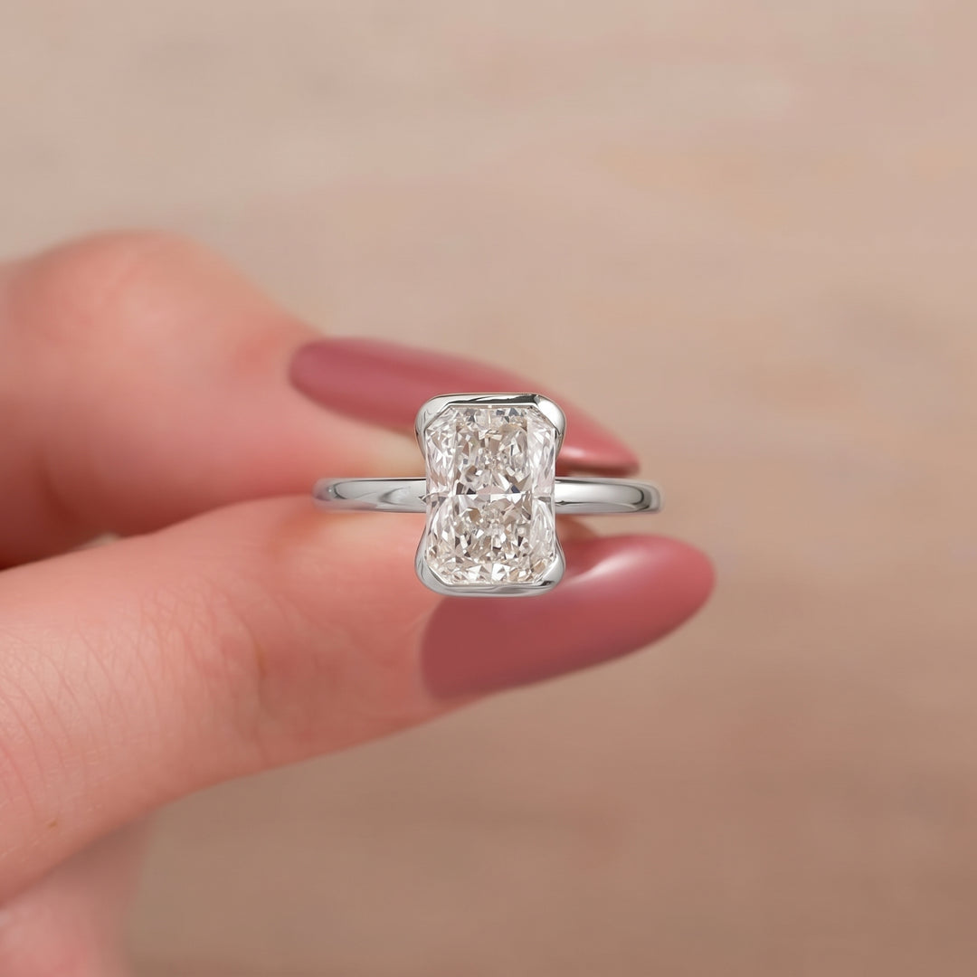 A silver ring with a rectangular baguette-cut diamond set on a slim silver band, held between fingers against a soft beige background.#material_white