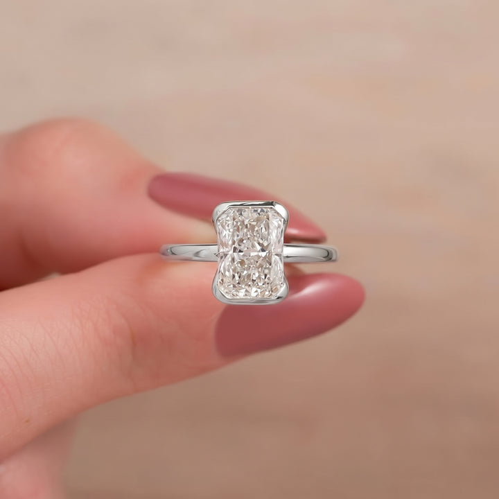 A silver ring with a rectangular baguette-cut diamond set on a slim silver band, held between fingers against a soft beige background.#material_white