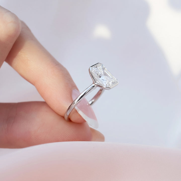 A silver ring with a rectangular emerald-cut clear gemstone held between a person's fingers against a soft, light pink and white background.#material_white