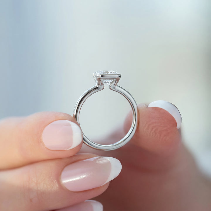 A close-up of a silver solitaire engagement ring held between fingers with a French manicure, set against a soft, out-of-focus light background.#material_white