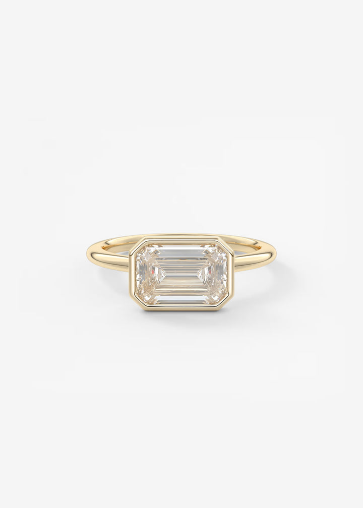 A gold ring featuring a large, bezel-set emerald-cut clear gemstone, photographed against a clean, light off-white background.#material_gold