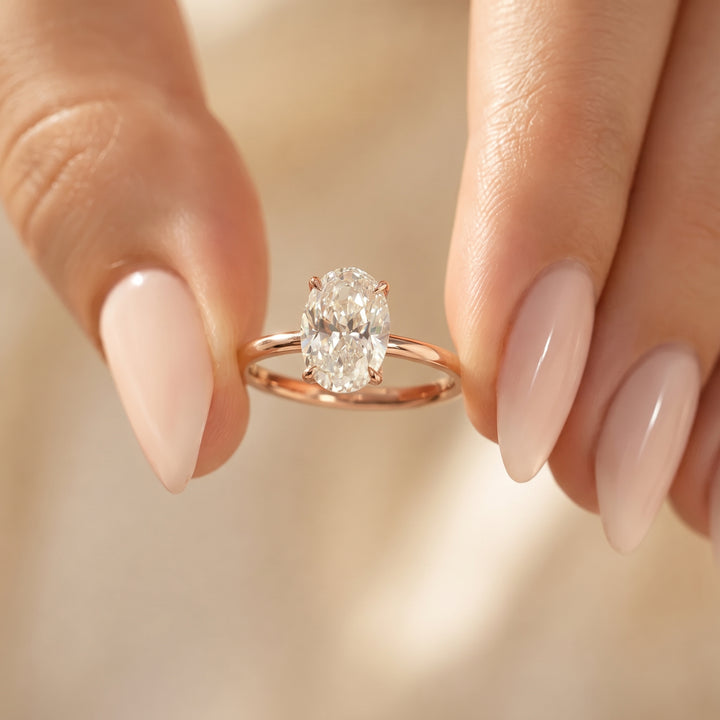 A close-up of a rose gold ring with an oval-cut clear gemstone held between two manicured fingers against a soft, warm beige background. #material_rose
