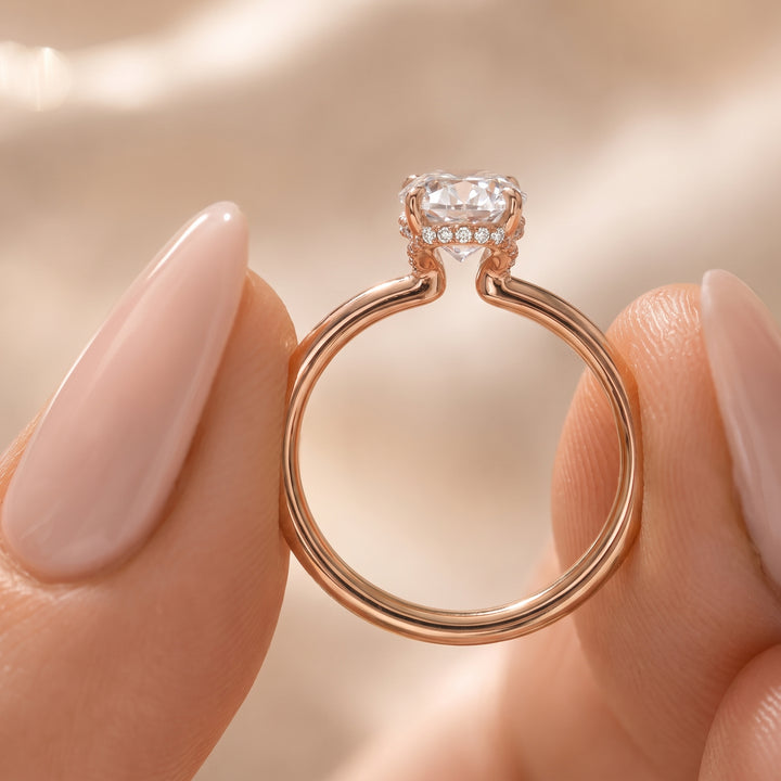 A rose gold ring with a heart-shaped clear gemstone held between two manicured fingers against a soft, warm beige background. #material_rose