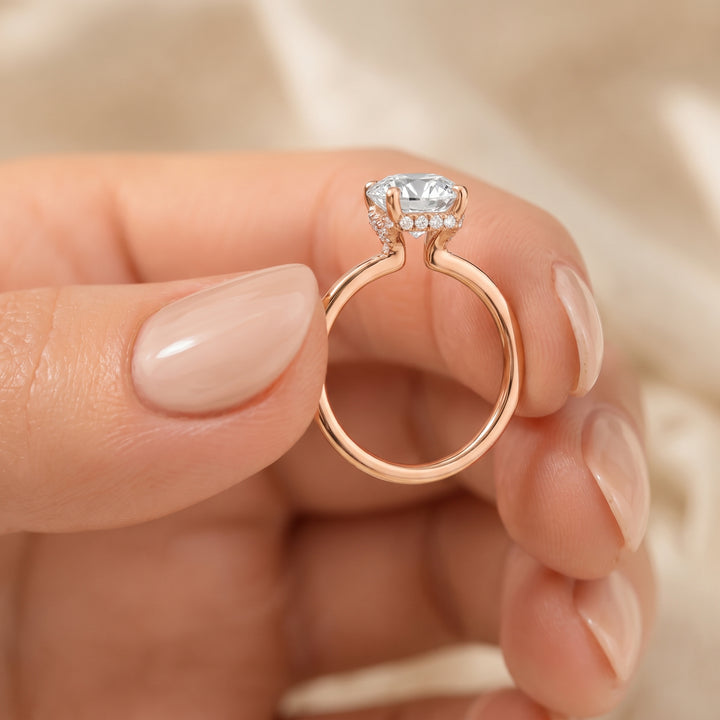 A close-up of a hand with manicured nails holding a rose gold ring with a large clear cushion-cut center stone and small accent stones. #material_rose
