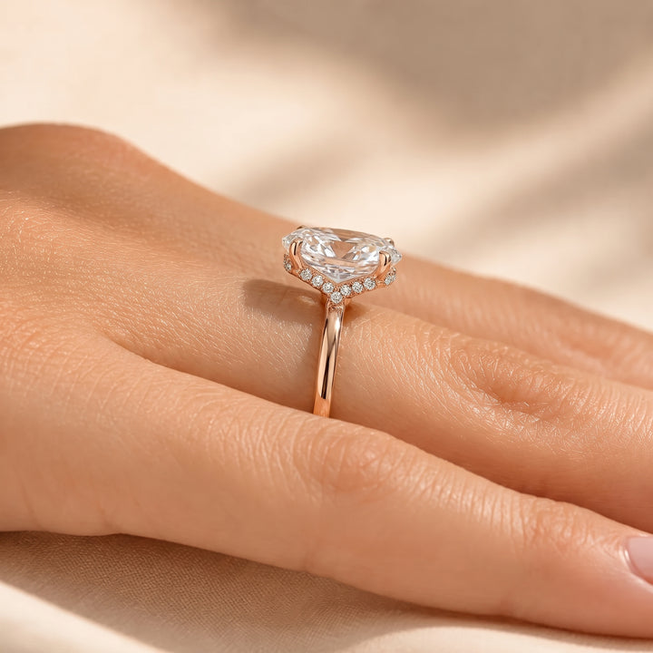 A close-up of a hand wearing a rose gold ring with a heart-shaped clear gemstone and small accent stones, set against a soft beige fabric background. #material_rose