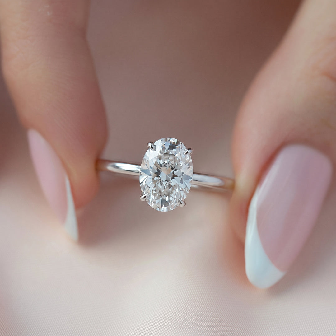 An oval-cut diamond solitaire ring on a polished silver band, held between two hands with pale pink and white French-tipped nails against a soft, light pink fabric background. #material_white