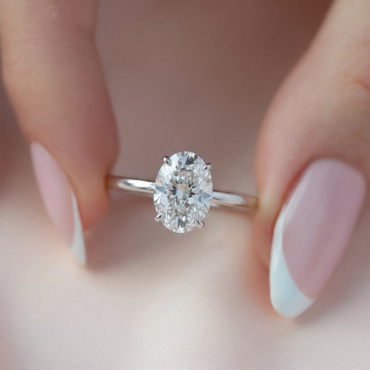 An oval-cut diamond solitaire ring on a polished silver band, held between two hands with pale pink and white French-tipped nails against a soft, light pink fabric background. #material_white