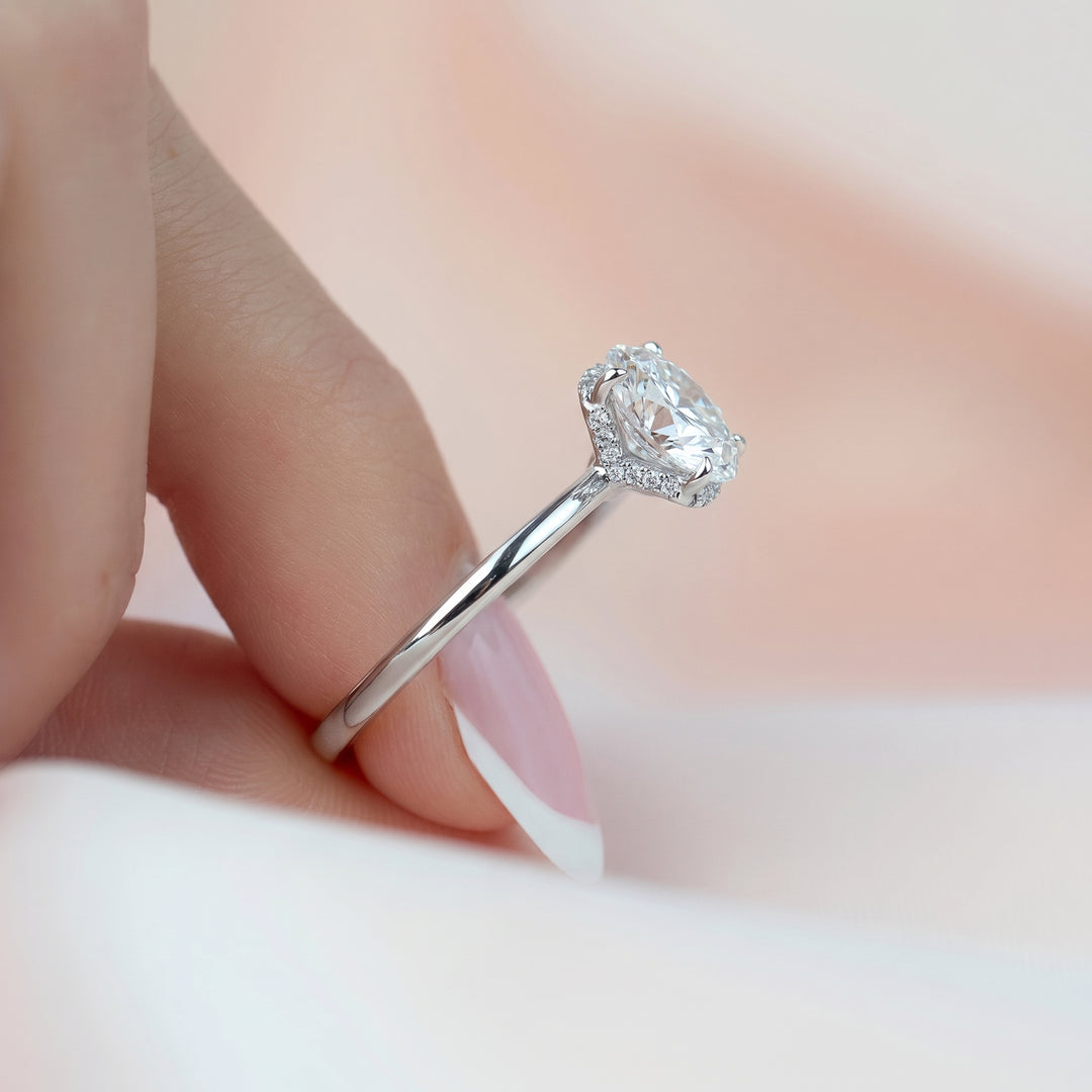 A close-up of a hand wearing a silver engagement ring with a pear-cut clear center stone and small accent stones, set against a soft, light pink and white background. #material_white