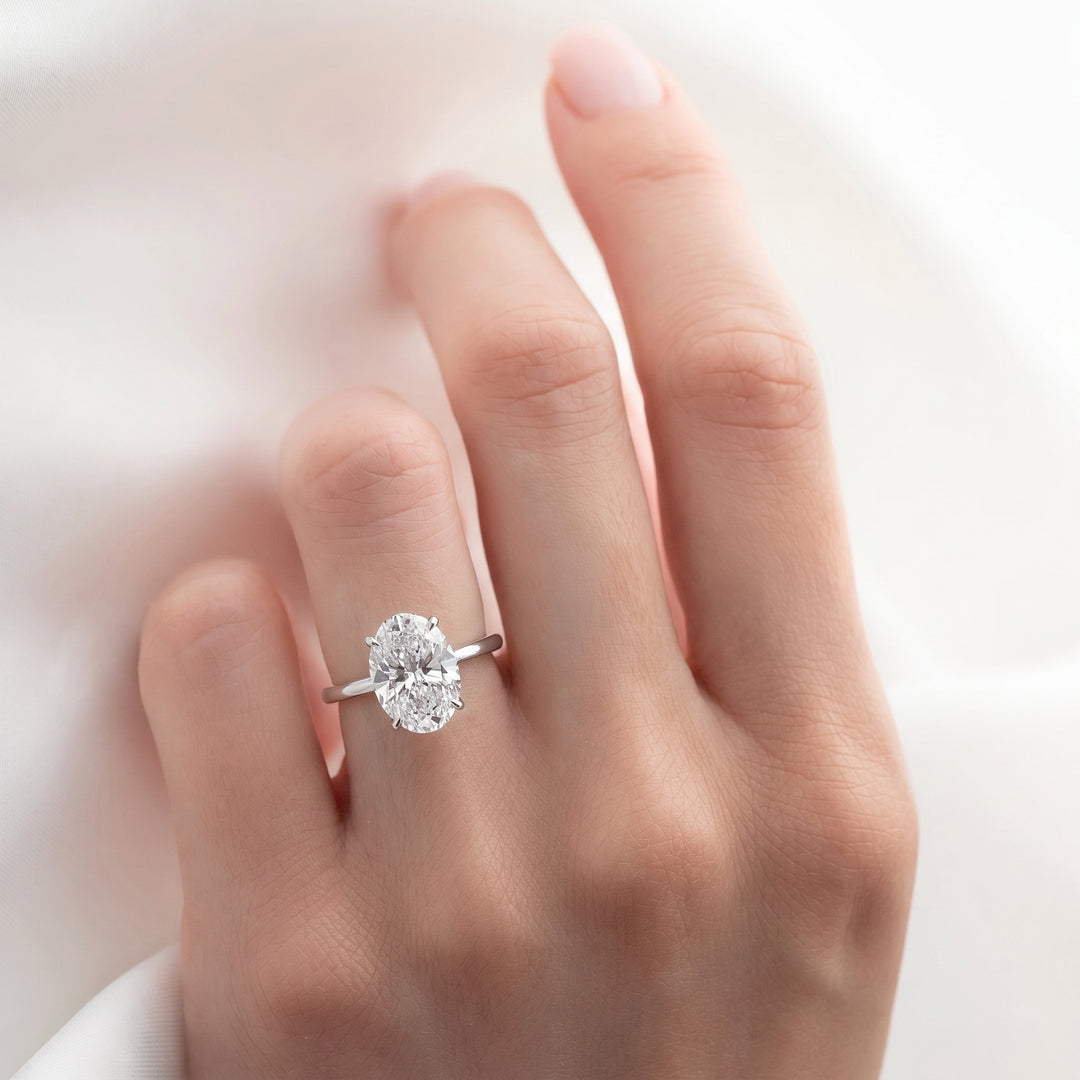 A close-up of a hand wearing a silver engagement ring with a round-cut clear gemstone, set against a soft white fabric background. #material_white