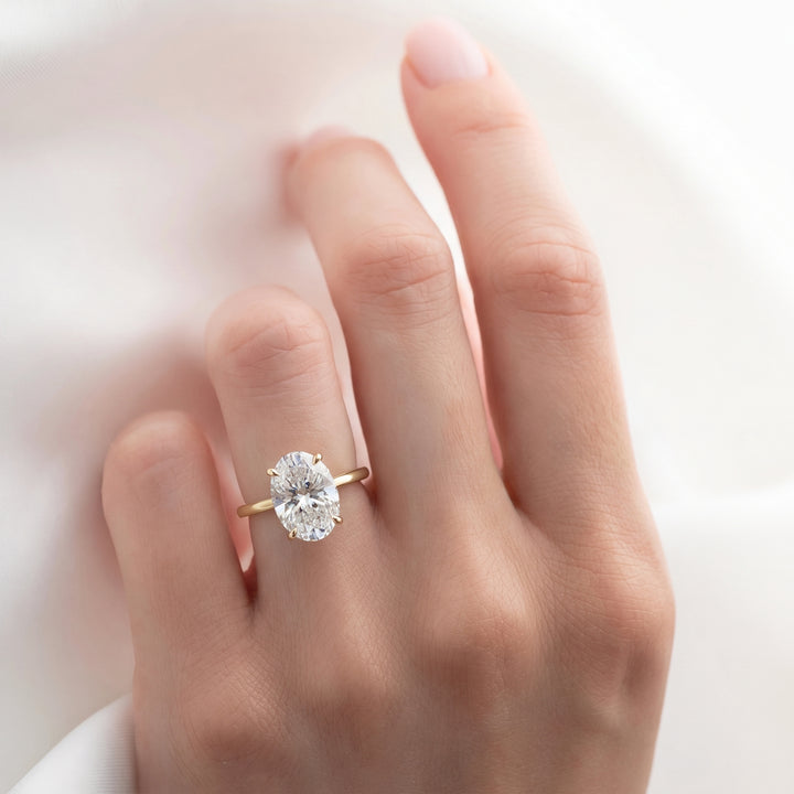 A close-up of a hand wearing a yellow gold ring with a large oval-cut clear gemstone, set against a soft white fabric background. #material_gold