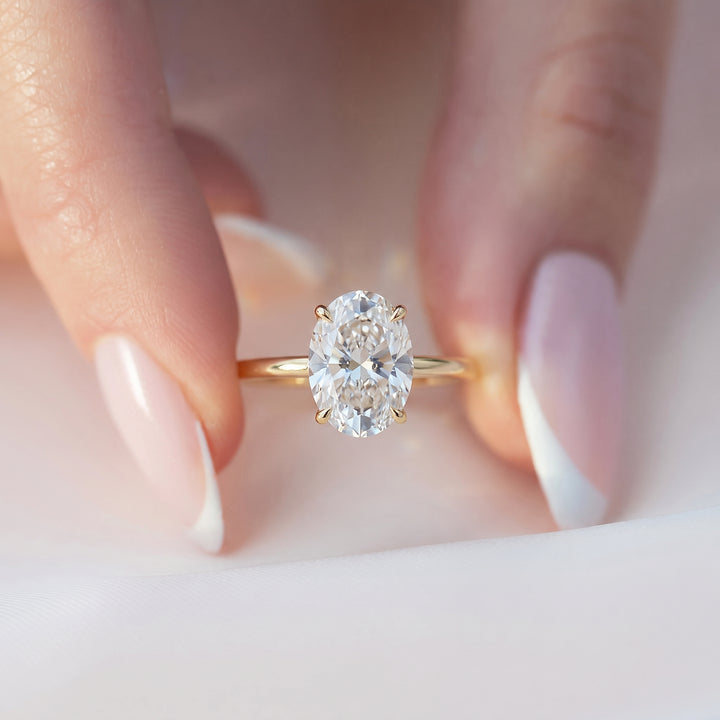 A close-up of a gold ring with a large oval-cut clear gemstone, worn on a person's finger with manicured nails. #material_gold