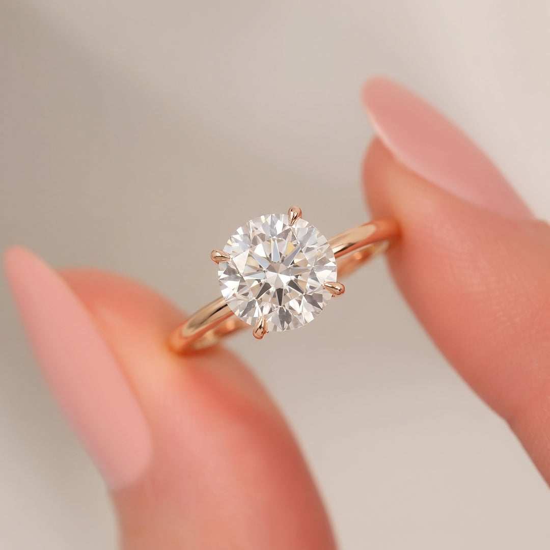 A round-cut diamond solitaire ring in a rose gold six-prong setting, shown being held between two fingers with manicured pink nails against a soft, light gray background. #material_rose