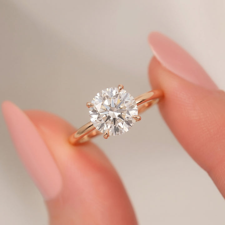 A round-cut diamond solitaire ring in a rose gold six-prong setting, shown being held between two fingers with manicured pink nails against a soft, light gray background. #material_rose