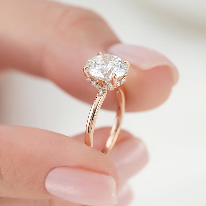 A close-up of a hand holding a rose gold engagement ring with a round-cut diamond center stone and small accent diamonds. #material_rose