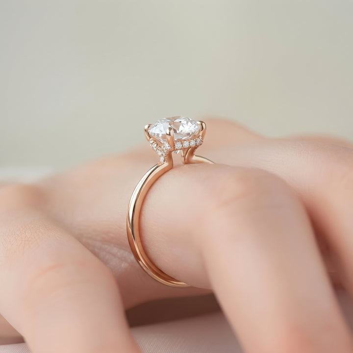 A rose gold engagement ring with a round-cut clear center stone and small accent stones, shown worn on a hand against a soft beige background. #material_rose