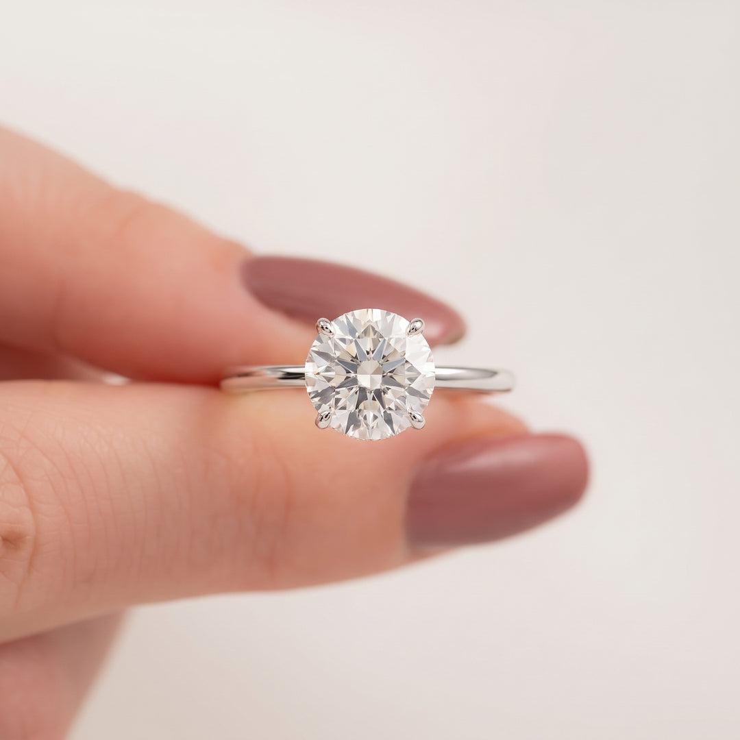 A classic solitaire diamond engagement ring with a round brilliant-cut center stone on a polished silver band, held between fingers with mauve-colored nail polish against a clean white background. #material_white