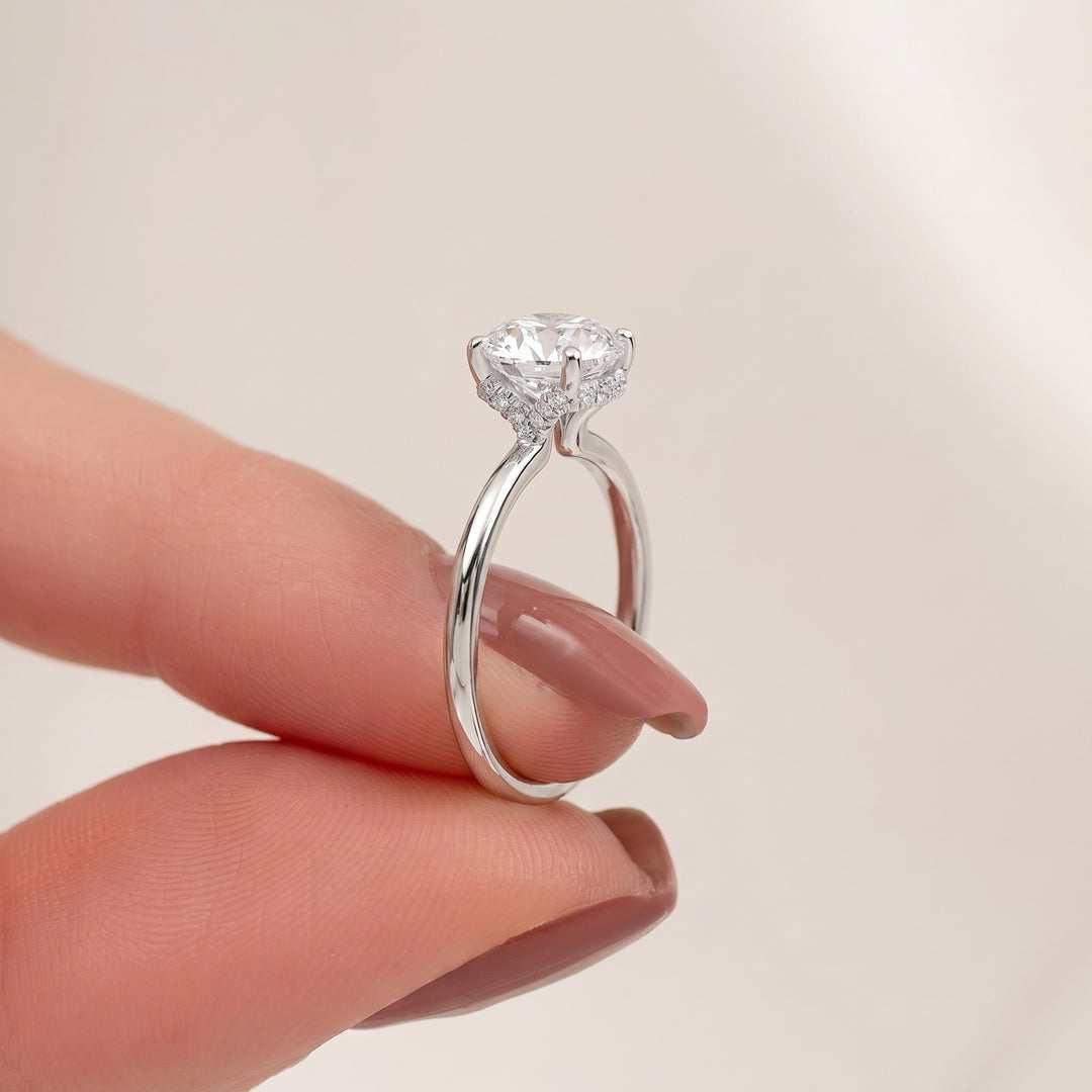 A silver solitaire engagement ring with a round brilliant-cut clear gemstone, held between two fingers against a soft, light beige background. #material_white