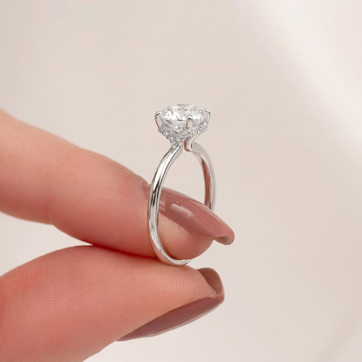 A silver solitaire engagement ring with a round brilliant-cut clear gemstone, held between two fingers against a soft, light beige background. #material_white