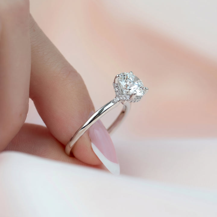 A close-up of a hand with a French manicure holding a silver engagement ring with a heart-shaped clear gemstone and a halo setting. #material_white