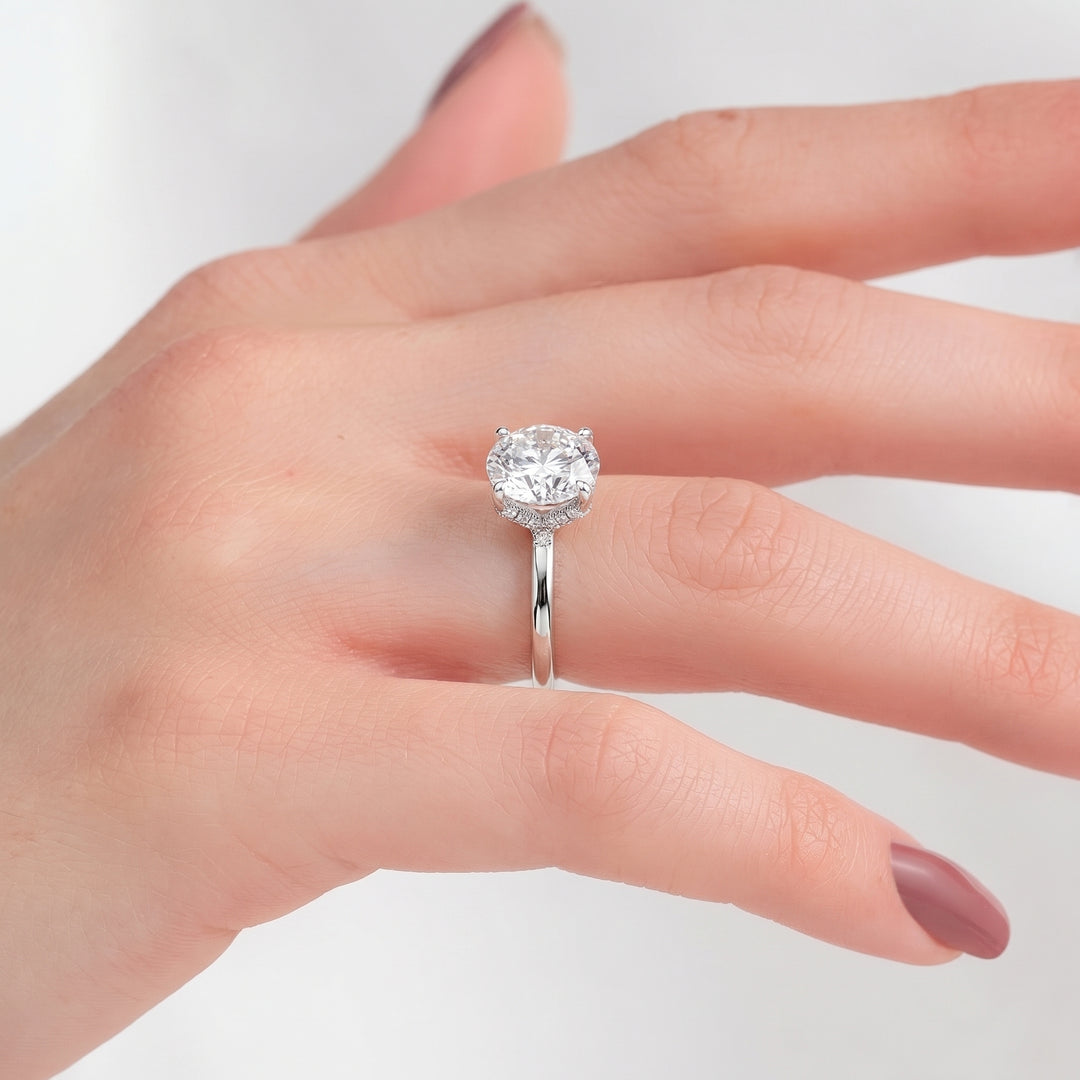 A silver engagement ring with a round-cut clear gemstone in a halo setting, shown worn on a hand with mauve nail polish against a light gray background. #material_white