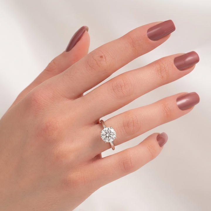 A close-up of a hand with mauve-colored nails wearing a classic solitaire engagement ring with a round-cut diamond on a rose gold band, set against a soft, light background. #material_white