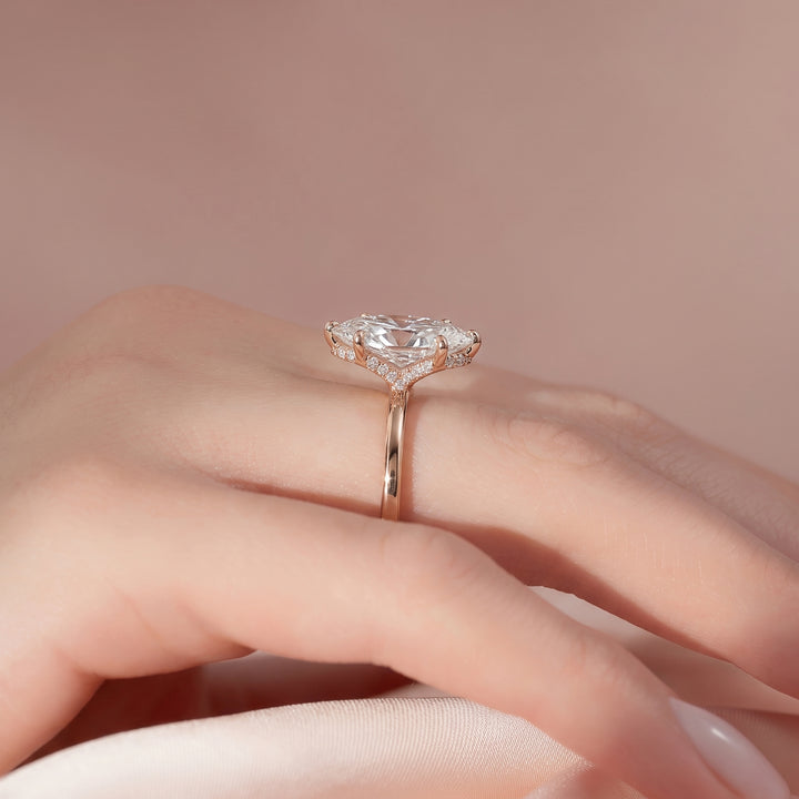 A close-up of a hand wearing a rose gold ring with a large heart-shaped clear gemstone and small accent stones, set against a soft pink background.#material_rose