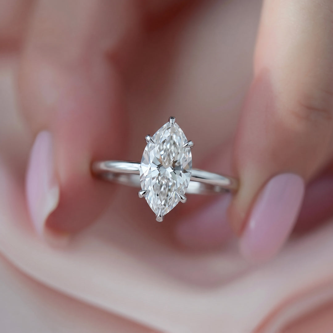 A close-up of a silver ring with a marquise-cut diamond solitaire, held between fingers against a soft pink background. #material_white