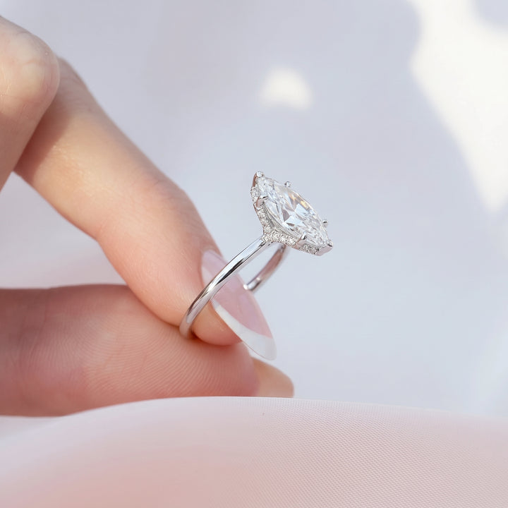 A silver-tone ring with a marquise-cut clear gemstone held between fingers against a soft white background. #material_white