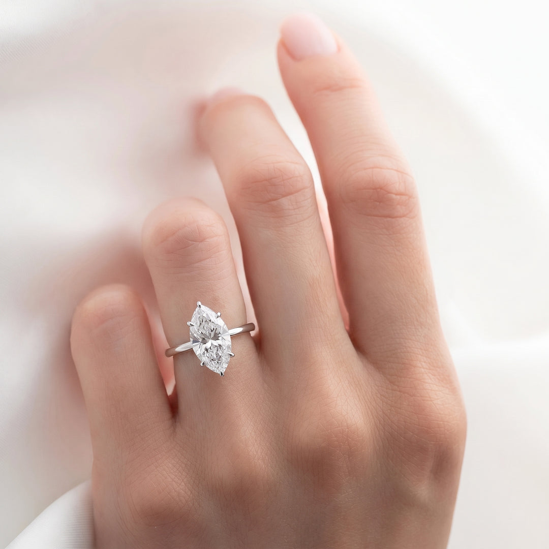 A close-up of a hand wearing a silver ring with a marquise-cut clear gemstone, set against a soft white fabric background.#material_white