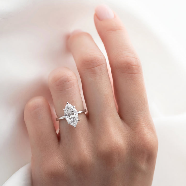 A close-up of a hand wearing a silver ring with a marquise-cut clear gemstone, set against a soft white fabric background.#material_white