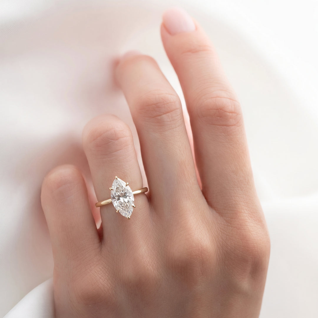 A close-up of a hand wearing a gold ring with a marquise-cut clear gemstone, set against a soft white background.#material_gold
