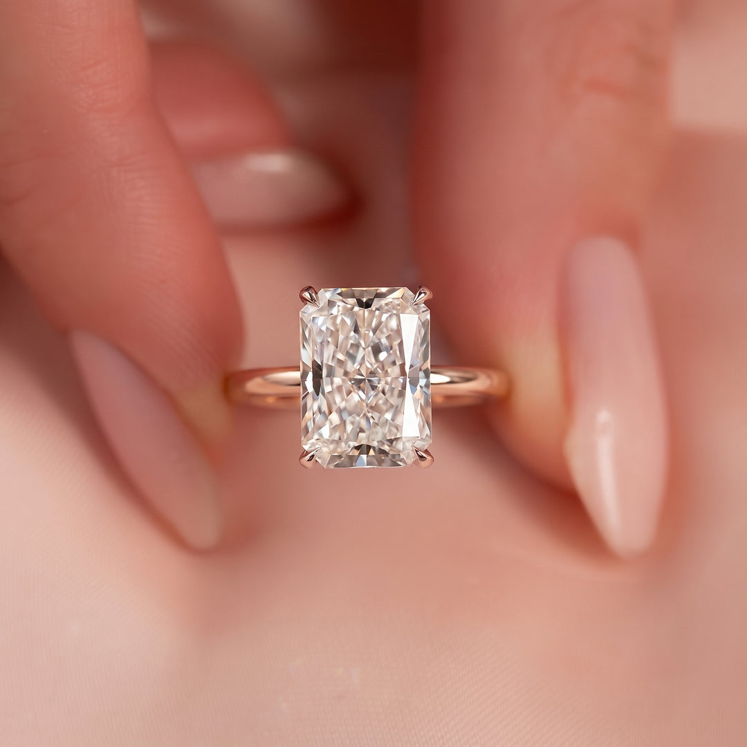 A close-up of a rose gold ring with a rectangular radiant-cut diamond, held between two fingers with manicured nails. #material_rose