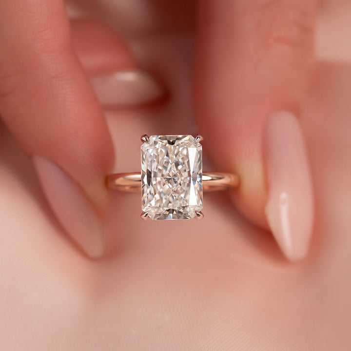 A close-up of a rose gold ring with a rectangular radiant-cut diamond, held between two fingers with manicured nails. #material_rose
