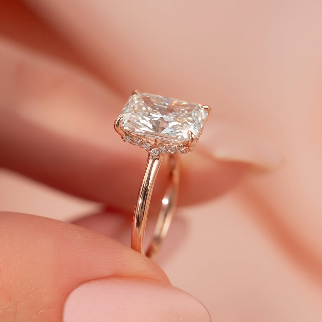 A close-up of a rose gold ring with a cushion-cut diamond and small accent diamonds, held between fingers against a soft pink background. #material_rose