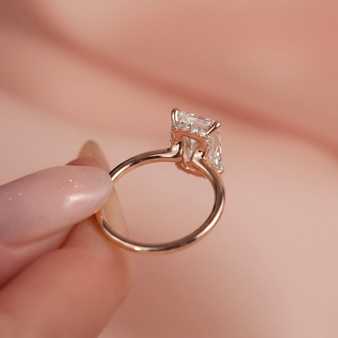 A close-up of a rose gold ring with a clear rectangular gemstone held by fingers against a soft pink background. #material_rose