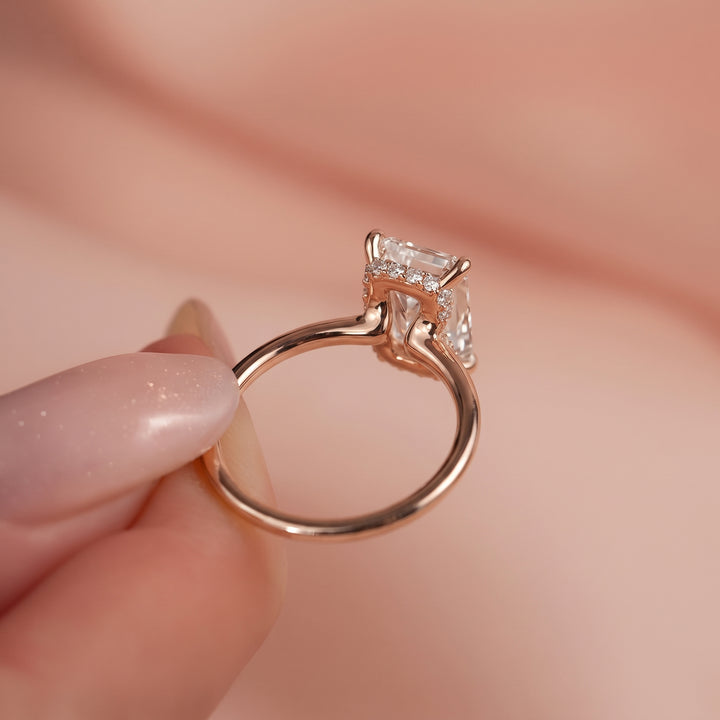 A close-up of a rose gold ring with a clear rectangular gemstone held by fingers against a soft pink background. #material_rose