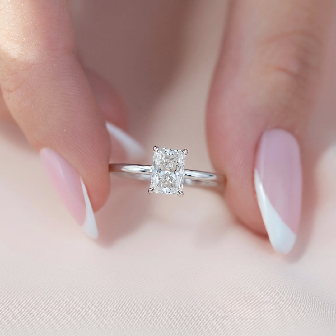 A close-up of a silver engagement ring with a princess-cut diamond center stone, held between two hands with French-tipped nails against a soft pink background. #material_white