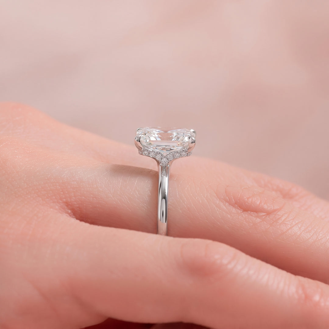 A close-up of a silver ring with a heart-shaped clear gemstone and small side stones, worn on a person's finger against a soft pink background. #material_white