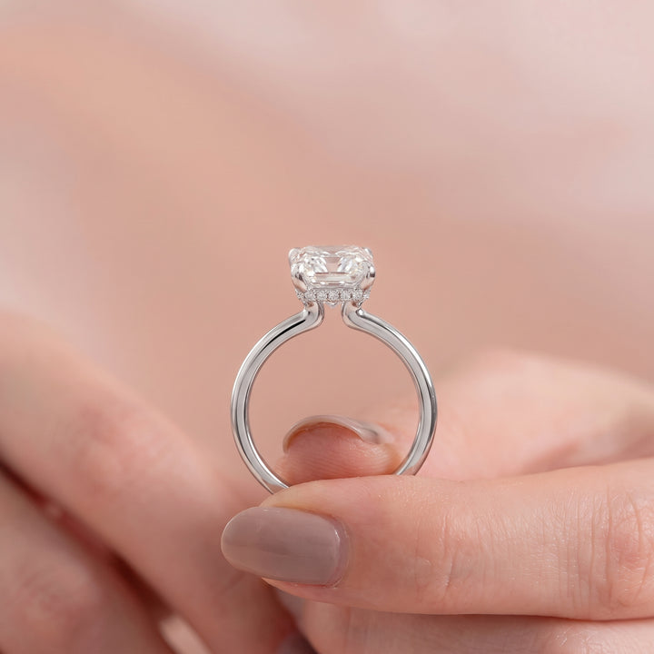 A silver solitaire engagement ring with a cushion-cut clear gemstone, held between two fingers against a soft, warm pink background. #material_white