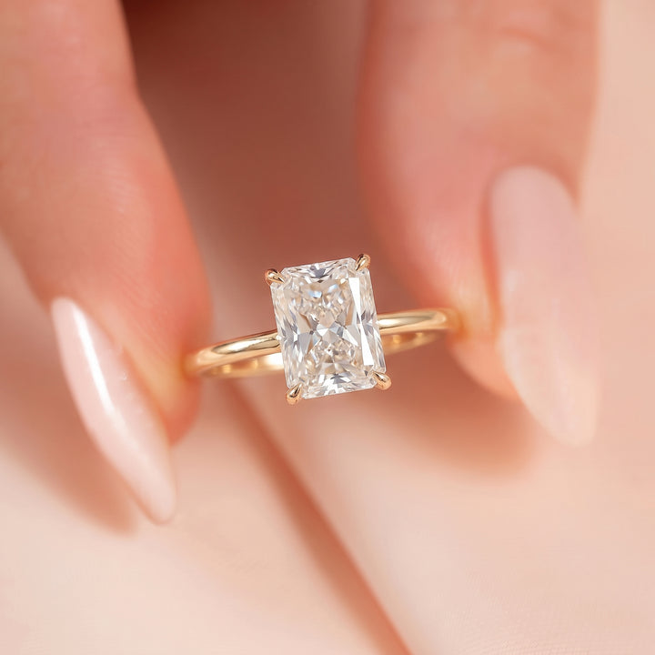 A close-up of a gold ring with a rectangular clear gemstone being held between fingers with pale pink manicured nails. #material_gold