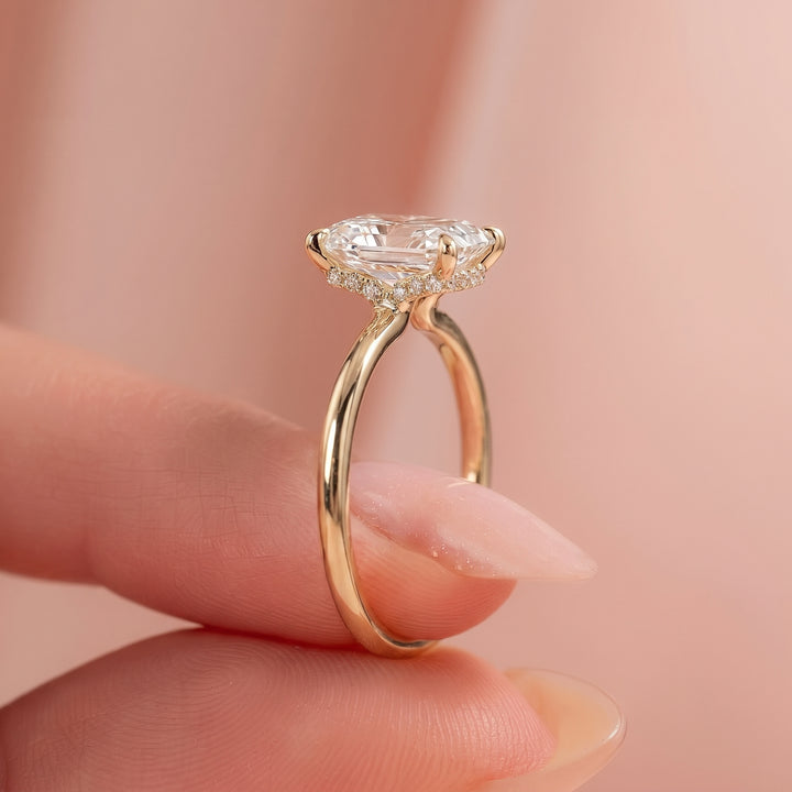 A close-up of a gold ring with a large clear heart-shaped gemstone and small accent stones, held between fingers against a soft pink background. #material_gold