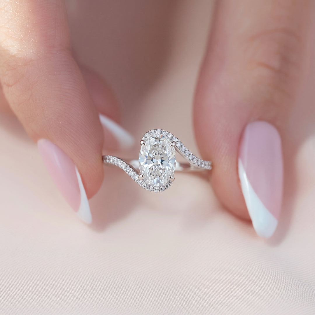 A close-up of a silver ring with a large oval-cut clear gemstone and smaller accent stones, shown being held between fingers with a soft pink French-tip manicure. #material_white