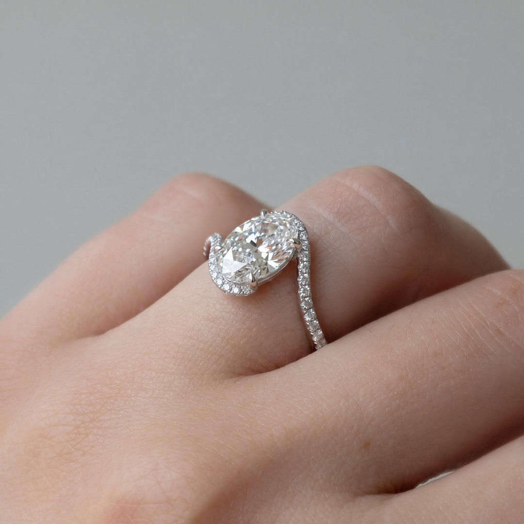 A close-up of a hand wearing a silver-toned ring with a large oval-cut clear center stone and a curved pavé band, set against a soft gray background. #material_white