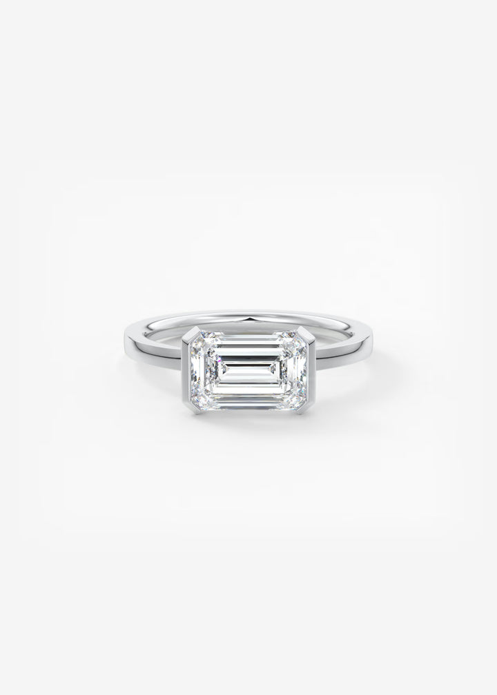 An emerald-cut diamond engagement ring in a silver-toned metal band, centered on a clean white background.#material_white